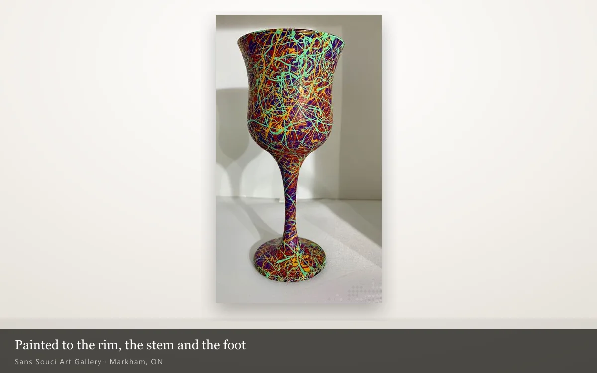 A hand-painted wine glass seen whole against a lit gallery wall, painted to the rim, down the stem and across the foot