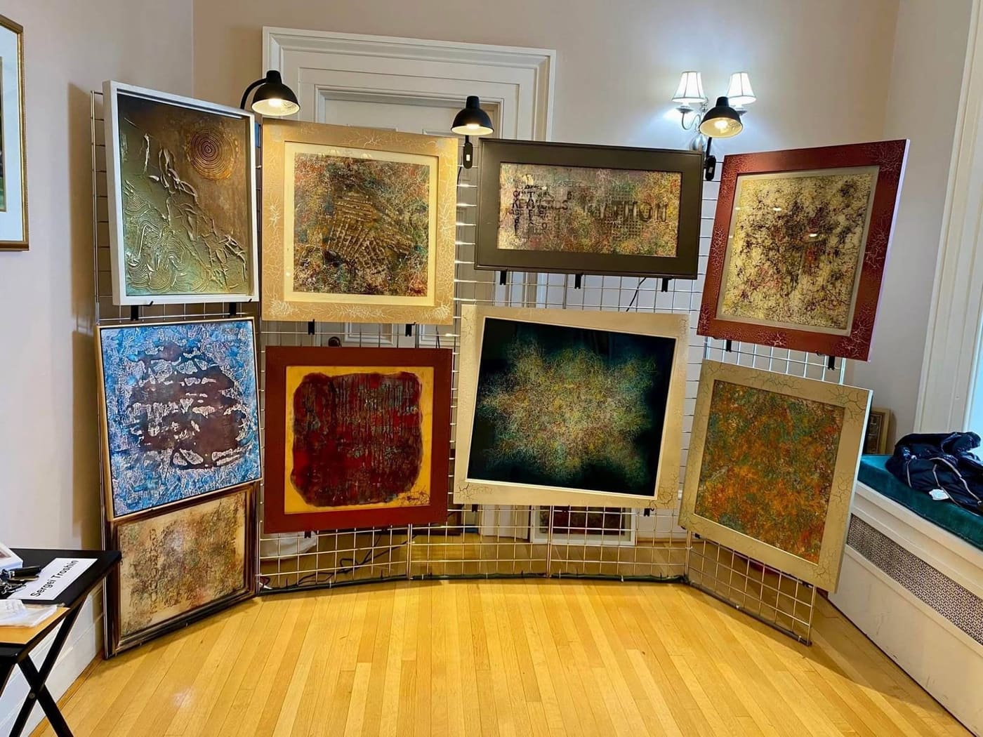 Nine framed paintings by Sergei Troshin on a curved white grid in a room with a white door, lamps and a window seat