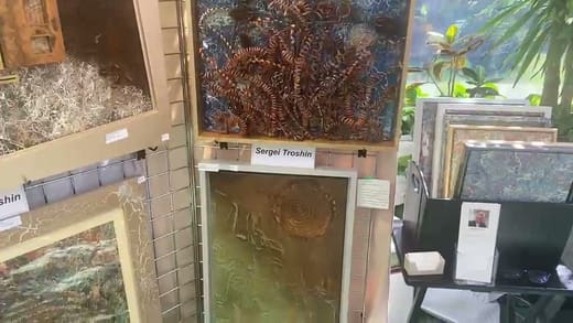 Still from the artist's video: a gold textured panel in a white frame below The Choice, beside a stand of prints