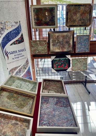 Looking down at the display: framed paintings on a black grid against a window, the Sans Souci banner on the left and framed pieces laid flat on a red table