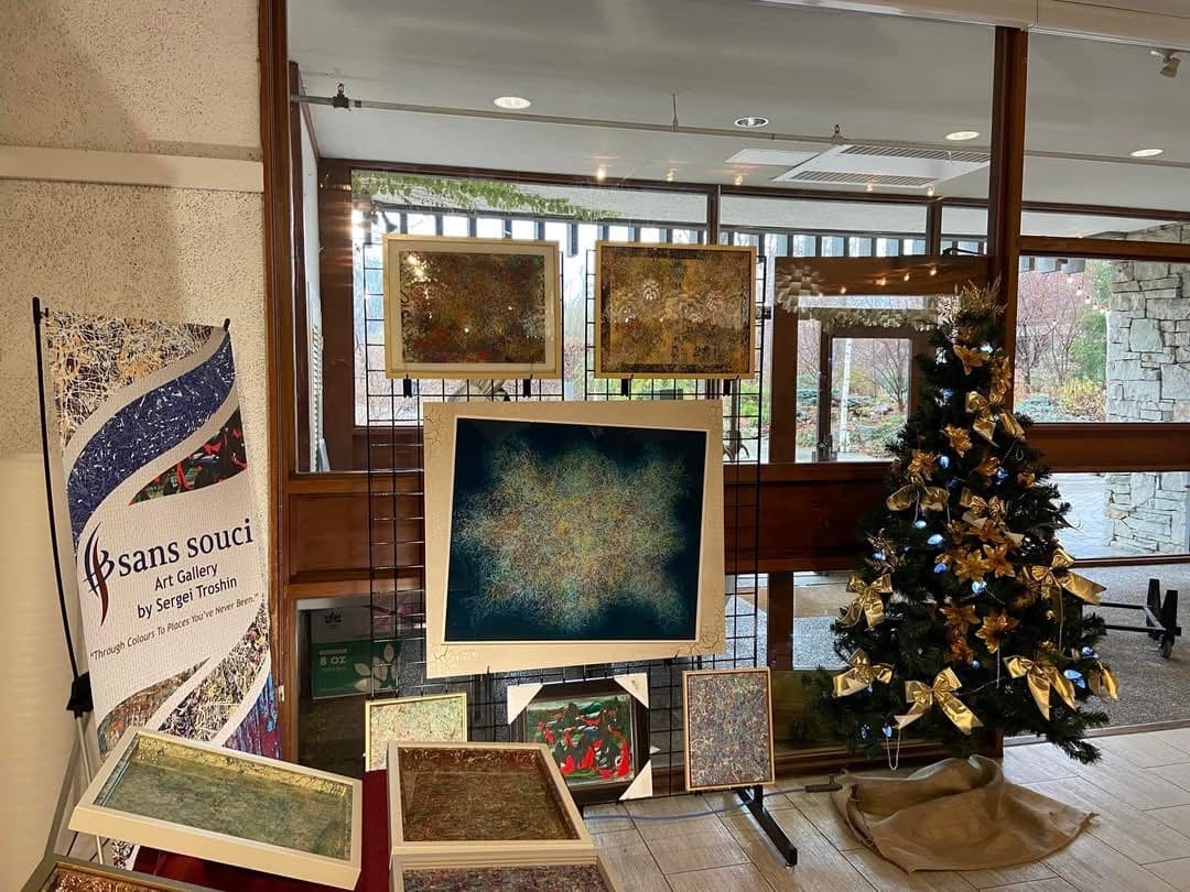 Sans Souci Art Gallery banner and framed paintings on a black grid by tall windows, next to a Christmas tree with gold bows