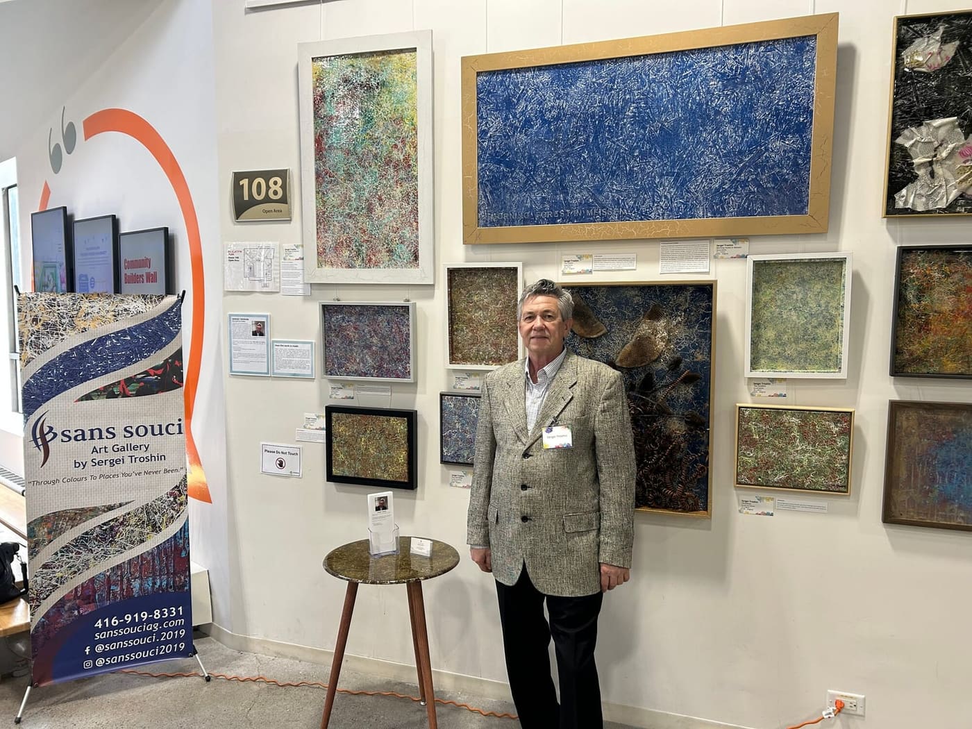 Sergei Troshin standing in front of his wall of framed paintings at 105 Gibson Centre, with the Sans Souci banner on the left