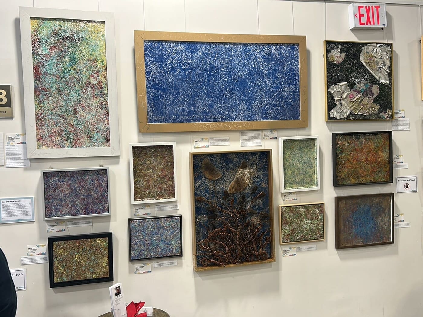 Wall of framed paintings by Sergei Troshin: a long blue panel in a gold frame at the top, a blue panel with metal spirals in the middle, and smaller framed works around them